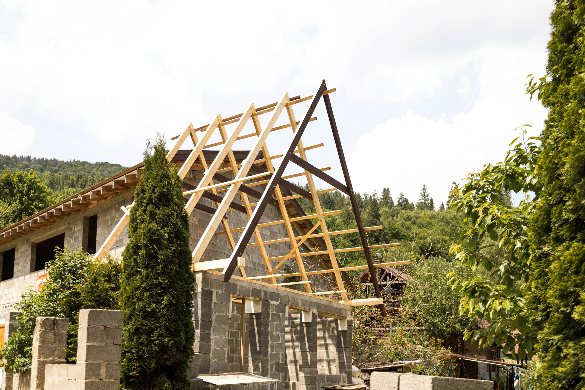 Ossature bois d'une extension de maison en cours de montage, accolée à la façade existante d'une habitation en pierre. Structure en madriers massifs traditionnels assemblés, toiture en tuiles terrain cuite partiellement posée, bardage bois clair à demi-installé. Charpente traditionnelle sans structure métallique visible. Chantier en plein air par beau temps.